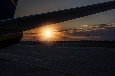 The image captures the sun gradually setting in the horizon, casting a warm glow behind the wing of a commercial airplane in flight. The silhouette of the aircraft contrasts against the vibrant colors