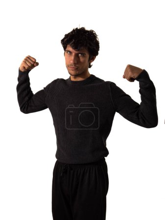 Ein Mann in schwarzem Pullover und schwarzer Hose lässt seine Muskeln in einer selbstbewussten Pose spielen. Der Kontrast der schwarzen Kleidung zu seinem durchtrainierten Körperbau verstärkt die optische Wirkung des Bildes.