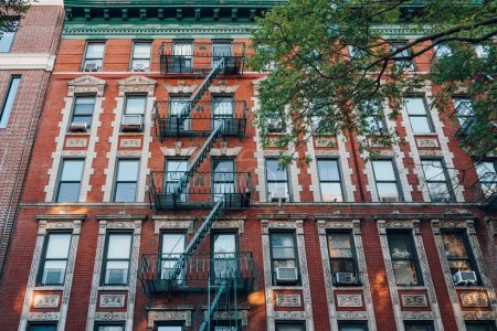 Fachada de un típico bloque de apartamentos de ladrillo rojo de Nueva York con escalera de incendios en la parte delantera, árbol en frente.