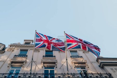 Foto de Vista de bajo ángulo de dos banderas británicas Union Jack que fluyen en el viento, en postes fuera de un edificio en Londres, Reino Unido. - Imagen libre de derechos