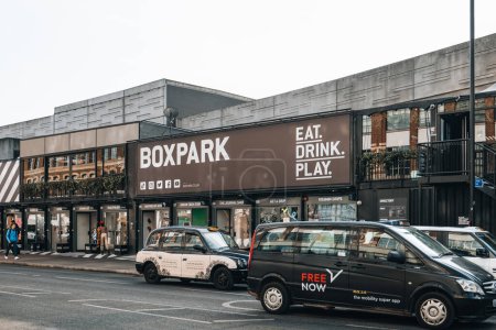 Photo for London, UK - February 09, 2023: Cars and taxi in front of shops at Boxpark Shoreditch, London, shipping container pop-up mall for independent and fashion and lifestyle stores and cafes. - Royalty Free Image