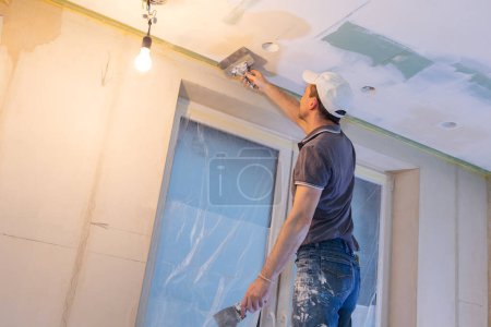 Téléchargez les photos : Travailleur faire des réparations dans un nouvel appartement. Homme plâtre murs et plafonds. Photo de haute qualité - en image libre de droit
