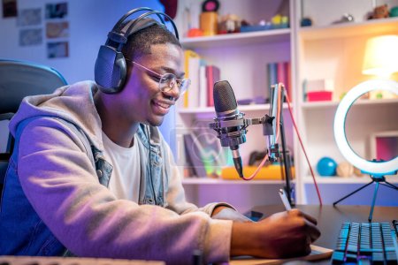 Young smiling man wearing headphones and talking into a microphone at the radio station recording podcast. Male sound producer working in home recording studio. Afroamerican man streamer playing video