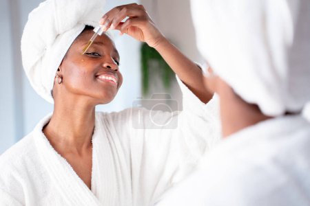 Photo for Beauty young woman applying serum vitamin c, argan oil tea tree in pippette dropper on cheek, treating skin for nourishing and moisturizing effect. Looking to a mirror white badroom High quality photo - Royalty Free Image