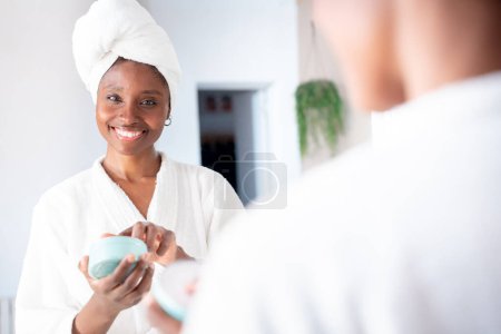 Foto de Retrato de mujer feliz aplicando crema cosmética natural, sonriendo mirando en el espejo. Cuidado de la piel rutina aseo a sí misma después de la ducha en el baño - Imagen libre de derechos