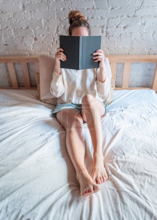 Foto de Tiempo perezoso en la cama. Mujer joven irreconocible relajarse por la mañana sentado en la cama sosteniendo y leyendo un libro en un acogedor dormitorio. Copiar espacio foto vertical. Foto de alta calidad - Imagen libre de derechos