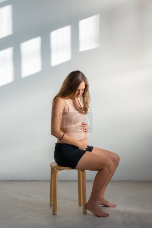 Téléchargez les photos : Joyeux jeune belle femme enceinte toucher et caresser son ventre et regarder vers le bas debout à la maison. Photo de haute qualité - en image libre de droit