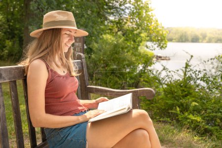 Foto de Mujer joven y relajada con un sombrero leyendo un libro sentado en un banco al aire libre en un día soleado. Tiempo libre en la naturaleza, foto tranquila frente a un lago. Foto de alta calidad - Imagen libre de derechos