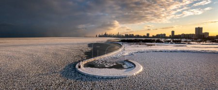Foto de Beautiful Chicago skyline aerial drone view from above the frozen ice and snow covering Lake Michigan with a curved hooked pier near Montrose beach with colorful orange and pink sunset clouds above. - Imagen libre de derechos