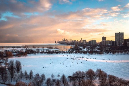 Foto de Crowds of people and families gather at Cricket hill to go sledding on a winter evening as the sunset begins to light up the clouds and sky in hues of yellow and pink. - Imagen libre de derechos