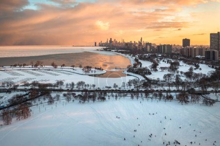 Beautiful aerial sunset photograph of Chicago skyline above cricket hill and Montrose Harbor with colorful pink and yellow clouds reflecting off the water in an opening in ice covering Lake Michigan.
