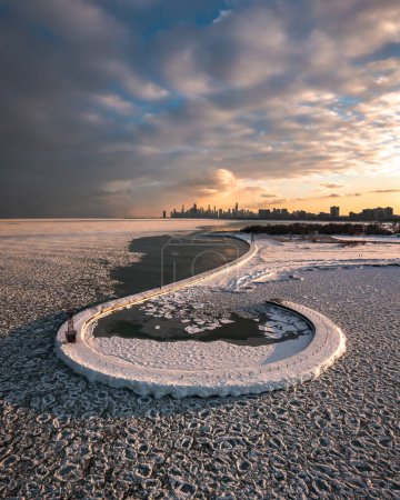 Photo for Beautiful aerial of a snow covered hook shaped pier surrounded by shards and round ice chunks floating on the cold water of Lake Michigan with the Chicago skyline beyond at sunset. - Royalty Free Image