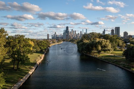 Téléchargez les photos : Aerial view of downtown Chicago skyline as a few people use the South Lagoon canal for standup paddle boarding, boating and rowing on a sunny day with blue sky and fluffy white cumulus clouds above. - en image libre de droit