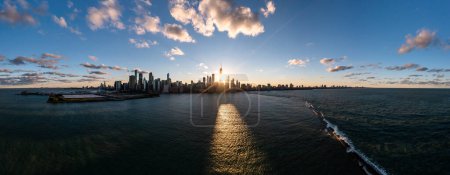 Foto de Wide angle aerial panorama of the Chicago skyline from Lake Michigan above the breakwater as the sun sets between highrise buildings casting a yellow glow on the water with blue sky and clouds above. - Imagen libre de derechos