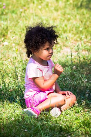 Photo for A cute young mixed race African American girl with dark brown curly hair and pink t-shirt and shorts sitting in shade on green grass while putting a piece of grass in her mouth on a sunny summer day. - Royalty Free Image