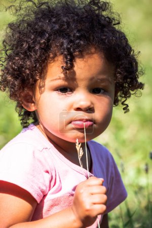 Téléchargez les photos : An adorable young toddler girl with dark brown curly hair sits on the grass on a sunny day staring into the distance as she puts a piece of grass in her mouth. - en image libre de droit