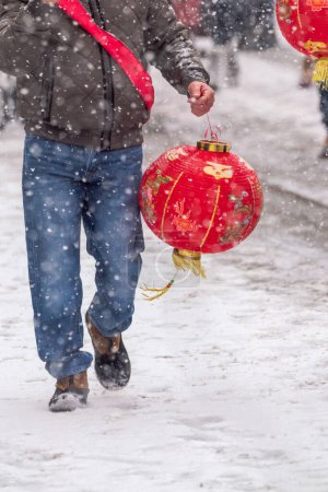 Foto de Chicago, IL - January 28th, 2023: A man in jeans and boots with a red cloth sash carries a festive red and gold Chinese lantern during the annual Argyle Lunar New Year parade in heavy snowfall. - Imagen libre de derechos