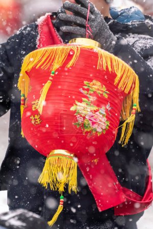 Foto de Chicago, IL - 28 de enero de 2023: Un hombre en un abrigo negro de invierno con una banda de tela roja lleva una linterna china festiva roja y dorada durante el desfile anual de Año Nuevo Lunar de Argyle en nevadas pesadas. - Imagen libre de derechos