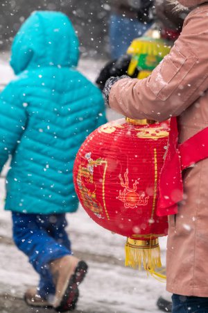 Téléchargez les photos : Chicago, IL - January 28th, 2023: A woman with a red cloth sash carries a festive red and gold Chinese lantern during the annual Argyle Lunar New Year parade in heavy snowfall. - en image libre de droit
