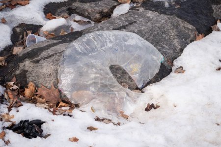 Foto de Close up of a partially deflated horseshoe or rainbow shaped clear balloon with colors mostly worn off and abandoned as litter along some rocks snow and leaves in winter. - Imagen libre de derechos