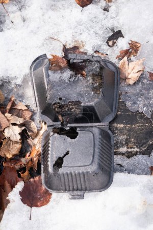 Photo for A close up of a torn up black extruded polystyrene sandwich box with ribs and holes laying on snow and leaves on top of limestone rock surface. - Royalty Free Image
