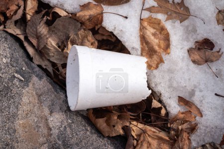 Foto de Una taza blanca de poliestireno extruido, hecha jirones y rota, con el fondo faltante, yace como basura entre rocas, hojas y nieve en temporada de invierno.. - Imagen libre de derechos