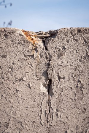 Téléchargez les photos : Close up of a crack in a concrete masonry wall with rust and rebar starting to show through with layers of beige or tan colored off white peeling paint and blue sky beyond. - en image libre de droit