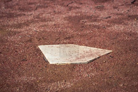 Téléchargez les photos : Close up of a white baseball home plate base in a field of wet brown or red colored dirt and mud on an empty baseball field on a sunny day. - en image libre de droit