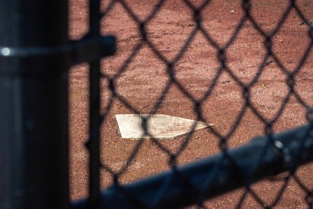 Foto de Close up of a white baseball home plate base from behind a black chain link fence in a field of wet brown and red colored dirt or mud on an empty or vacant baseball field on a sunny day. - Imagen libre de derechos