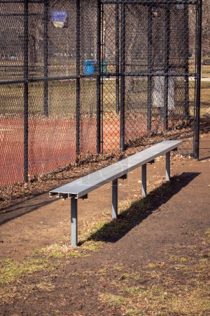 Foto de Primer plano de un banco de béisbol de metal de aluminio vacío en un campo de hierba y tierra con cerca de eslabón de cadena negro y campo de color rojo fangoso más allá. - Imagen libre de derechos