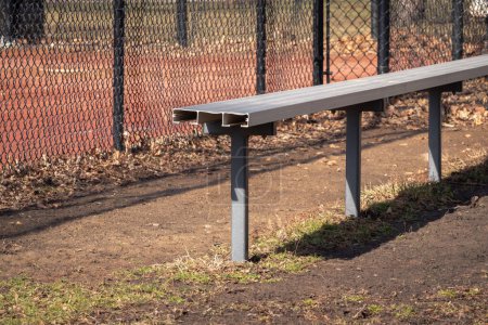 Foto de Close up of an empty aluminum metal baseball bench in a grass and dirt dugout with black chain link fence and muddy red colored infield beyond. - Imagen libre de derechos