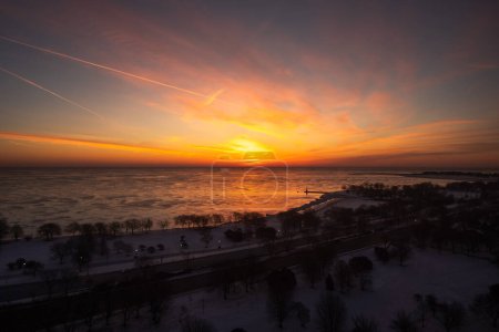 Téléchargez les photos : Aerial view of a colorful yellow orange and pink sunrise over Lake Michigan in winter with pieces of ice reflecting the orange glow on the water and snow on the ground between trees. - en image libre de droit