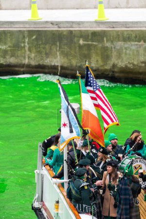 Téléchargez les photos : Chicago, IL - 11 mars 2023 : Un drapeau américain, Chicago et irlandais bat pavillon dans le vent au-dessus d'une bande celtique à bord d'un bateau avec d'autres passagers lors de l'événement annuel de teinture de la rivière verte au centre-ville. - en image libre de droit
