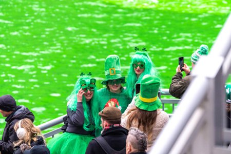 Téléchargez les photos : Chicago, IL - 11 mars 2023 : Un groupe de personnes vêtues de la tenue de la Saint-Patrick, y compris des cheveux verts et des chapeaux de lutin posent pour une photo devant la rivière verte fraîchement teinte au centre-ville. - en image libre de droit