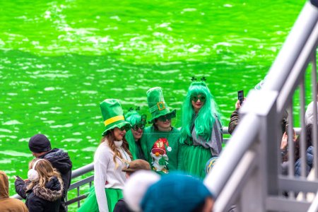 Téléchargez les photos : Chicago, IL - 11 mars 2023 : Un groupe de personnes vêtues de la tenue de la Saint-Patrick, y compris des cheveux verts et des chapeaux de lutin posent pour une photo devant la rivière verte fraîchement teinte au centre-ville. - en image libre de droit