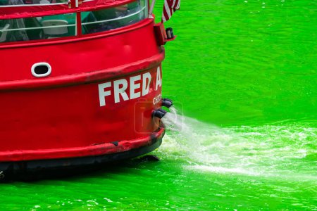 Foto de Chicago, IL - 11 de marzo de 2023: El barco Fred A. Busse, de color rojo brillante, navega por el río en contraste con el agua verde brillante del evento anual de teñido del río St. Patrick. - Imagen libre de derechos