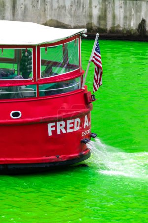Foto de Chicago, IL - 11 de marzo de 2023: Una bandera estadounidense cuelga de la parte posterior del barco Fred A. Busse rojo brillante que contrasta con el agua verde del evento anual de teñido del río St. Patrick 's Day. - Imagen libre de derechos