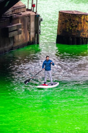 Téléchargez les photos : Chicago, IL - 11 mars 2023 : Un homme pagaie une planche à pagaie debout sous le pont DuSable sur la rivière verte fraîchement teinte lors de l'événement annuel de la Saint-Patrick. - en image libre de droit