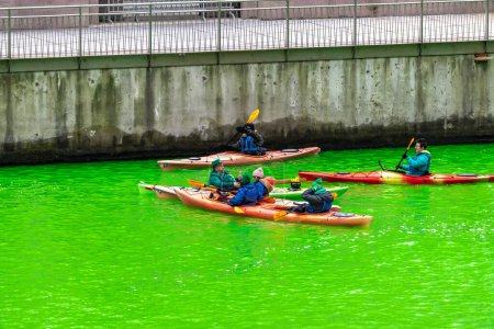 Photo for Chicago, IL - March 11th, 2023: A man dressed as a leprechaun with pipe in his mouth checks his phone while floating on the freshly dyed green water in a kayak during the annual St. Paddy's day event. - Royalty Free Image