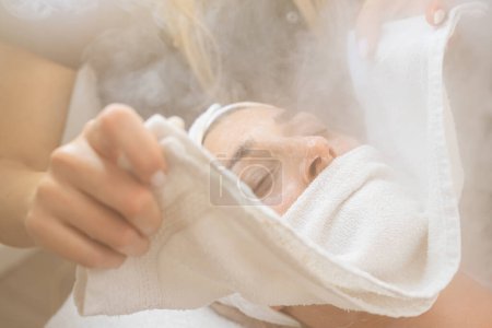 Nahaufnahme einer Frau mit geschlossenen Augen bei einer Wärmedampfbehandlung im Gesicht mit Handtuch in einem Kosmetiksalon oder Wellnessbereich. 