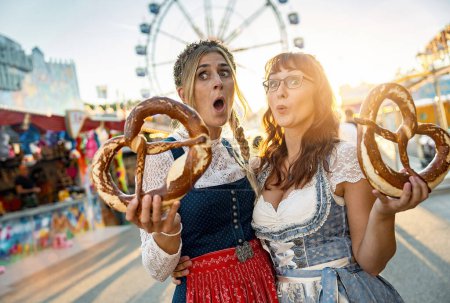 Téléchargez les photos : Deux petites amies heureuses ensemble sur une foire bavaroise ou oktoberfest ou terne en costume national ou Dirndl avec bretzel ou brezen en Allemagne - en image libre de droit