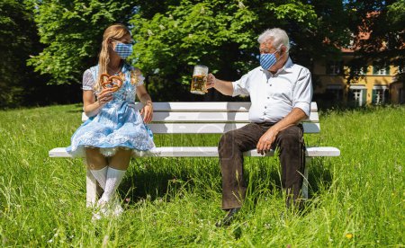 Téléchargez les photos : Distance sociale sur un banc en Bavière dans le jardin de la bière, senior et femme assis à part avec de la bière et bretzel - en image libre de droit