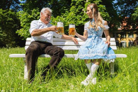 Téléchargez les photos : Senior et jeune femme assise sur un banc dans un jardin de bière bavarois ou oktoberfest toasting avec des tasses à bière allemandes - en image libre de droit