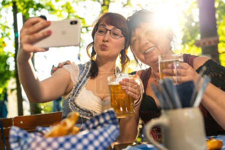 Photo for Best friends mother and daughter in Bavarian Tracht making a Selfie with the phone, with mugs of beer in Bavarian beer garden or oktoberfest - Royalty Free Image