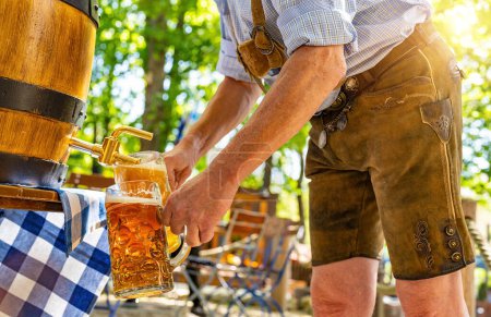Bavarian man in traditional leather trousers is pouring large lager beers in tap from wooden beer barrel in the beer garden. Background for Oktoberfest or Wiesn, folk or beer festival 