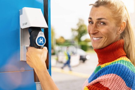 Foto de Hermosa mujer sonriendo, sostiene un dispensador de combustible con el logotipo de hidrógeno en la gasolinera para llenar su coche. motor de combustión h2 para la imagen concepto de transporte ecológico libre de emisiones - Imagen libre de derechos