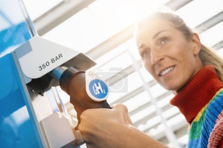 Photo for Beautiful woman smiling and looking away, holds a fuel dispenser with hydrogen logo on gas station to fill up her car. h2 combustion engine for emission free eco friendly transport concept image - Royalty Free Image