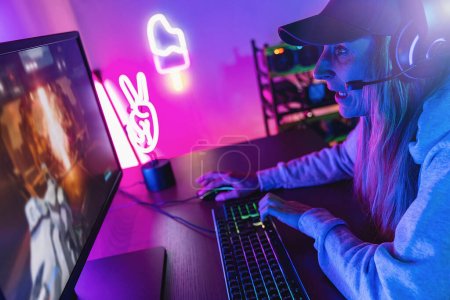 Photo for Agressive and shouting female Gamer Playing First-Person Shooter Online Video Game on her Powerful Personal Computer. Room and PC have Colorful Neon Led Lights. Young woman is Wearing a Cap at Home - Royalty Free Image