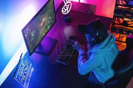 Photo for Professional female Gamer Playing First-Person Shooter Online Video Game on her Powerful Personal Computer. Room and PC have Colorful Neon Led Lights. Young Woman is Wearing a Cap at Home. - Royalty Free Image