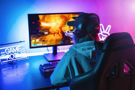 Chica jugando en primera persona Shooter Juego de vídeo en línea en su potente ordenador personal. La habitación y la PC tienen luces led de neón coloridas. Mujer joven está jugando con una gorra en casa